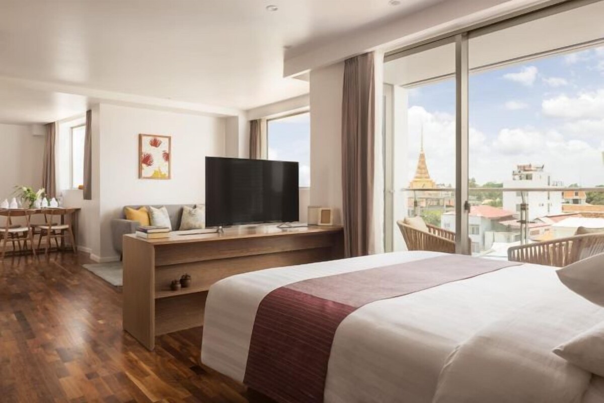 A hotel room with a bed, a TV on a wooden stand, a desk with a chair, and a large window with a view of buildings and a temple.