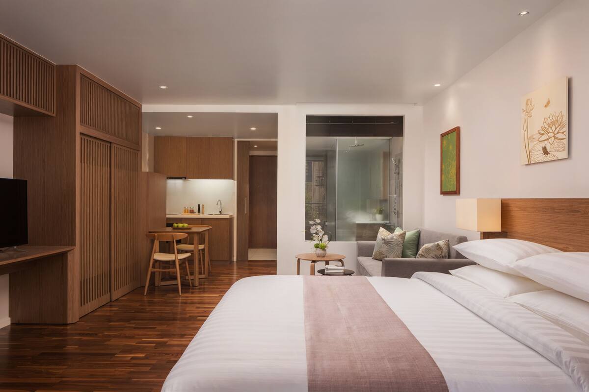 A modern hotel room with a large bed, a desk, a kitchenette, and a bathroom visible through a glass door.