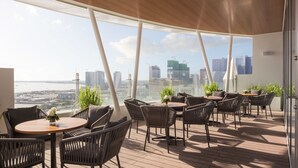 A modern outdoor seating area with a city view, featuring round tables and wicker chairs. - Hotel Emion Phnom Penh (Phnom Penh)