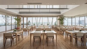 A spacious dining area with multiple tables and chairs, large windows offering a view of the cityscape, and potted plants adding greenery. - Hotel Emion Phnom Penh (Phnom Penh)