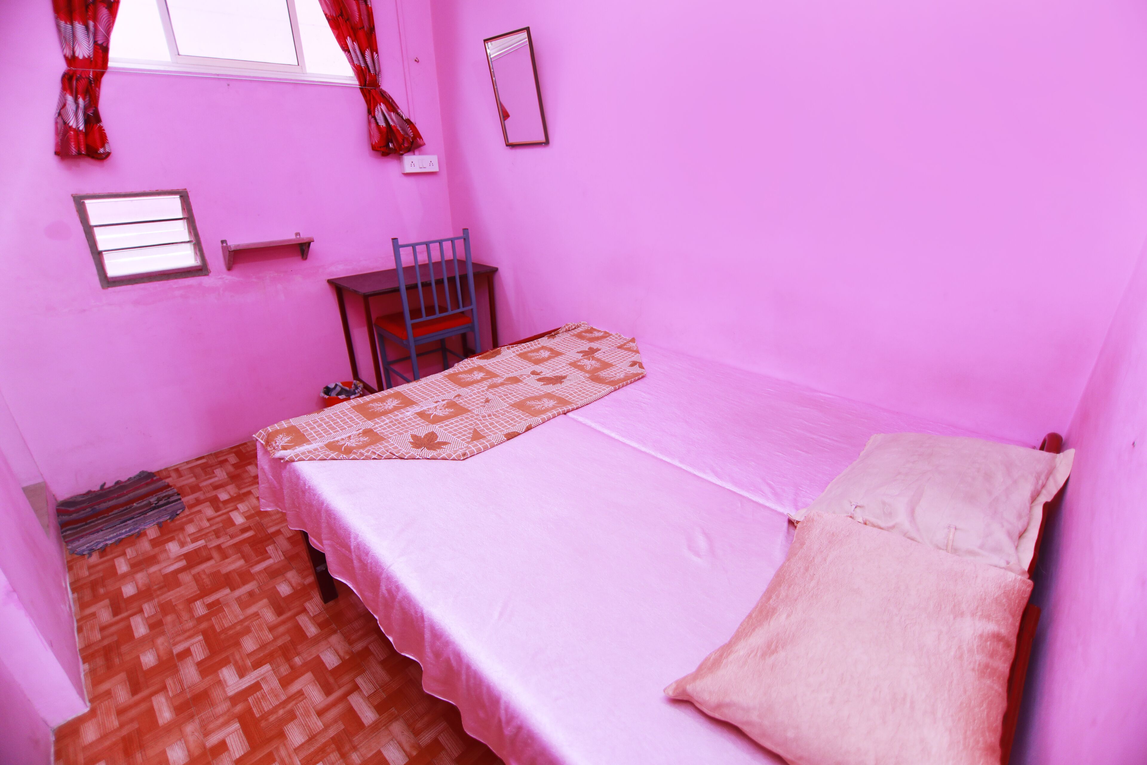 A small, pink-themed room with a single bed, a chair, a small table, and a window with curtains.
