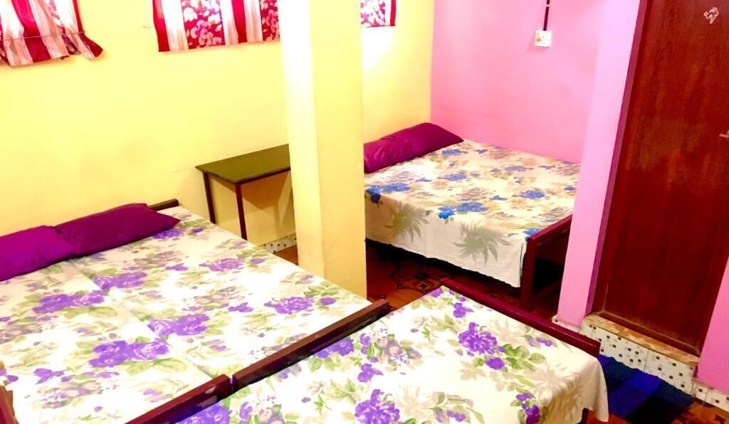 A small room with two beds, each with a floral bedspread and purple pillows. There is a small table and a wooden door.