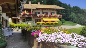 Um chalé tradicional com varanda decorada com flores, um caminho de pedra que leva à entrada e um jardim exuberante ao fundo.