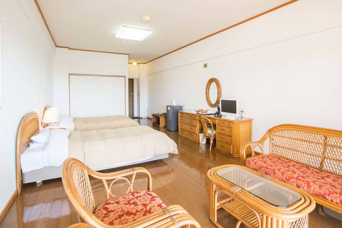 Une chambre spacieuse avec un grand lit, une commode, un bureau avec un ordinateur et deux fauteuils en osier.