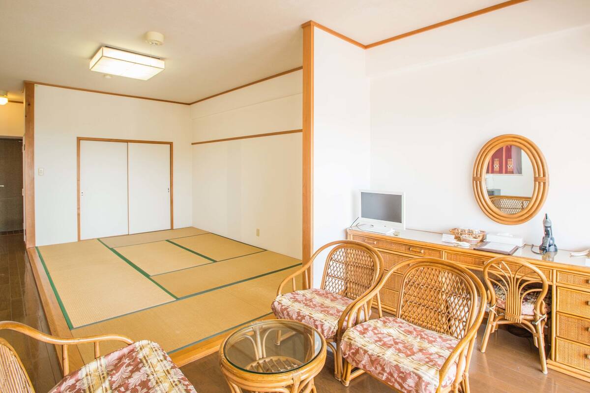 Une pièce avec un sol recouvert de tatamis, un bureau avec un ordinateur, des chaises en osier, un miroir et une porte coulissante traditionnelle japonaise.
