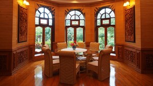 Un comedor con una mesa redonda, sillas de mimbre y grandes ventanales con detalles de madera tallada.