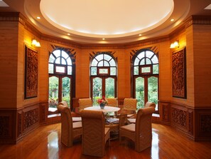 Relax in a serene space with a round table and wicker chairs, surrounded by intricate paneling and large windows with views of the lush surroundings.