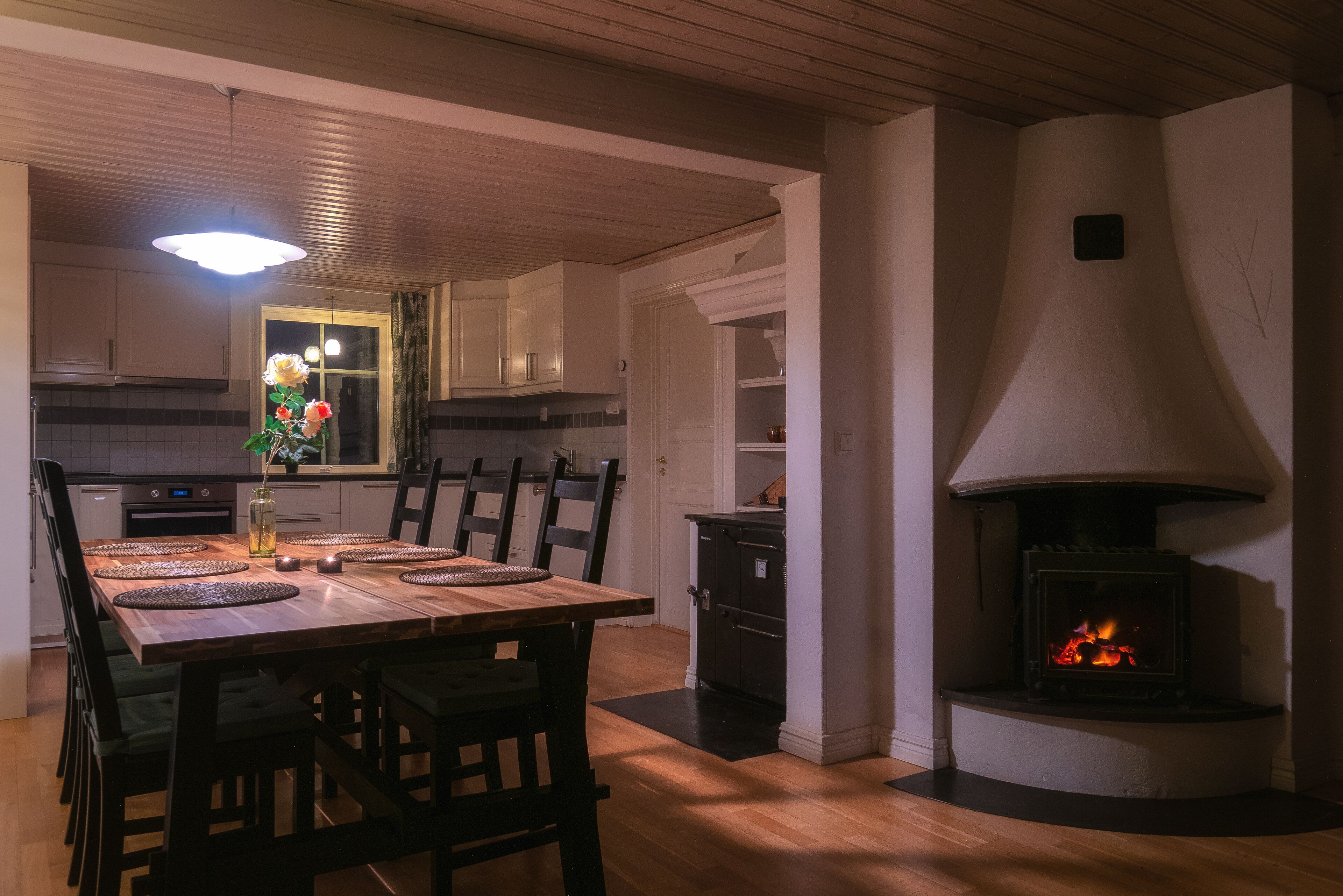 A dining area with a wooden table and chairs, a vase with flowers, and a fireplace with a visible fire.