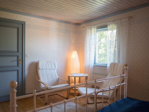Una habitación con techo de madera, una cama con cabecera blanca, una silla blanca, una mesita con lámpara y una ventana con cortinas.