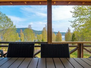 Una terraza de madera con sillas de mimbre y una mesa, con vistas a un lago y colinas boscosas.