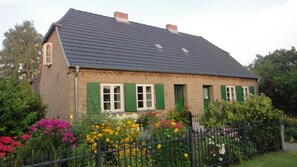 A two-story house with a dark tiled roof and green shutters, surrounded by a variety of colorful flowers and a black metal fence.