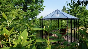 A glass gazebo with a metal frame, surrounded by lush greenery and trees.