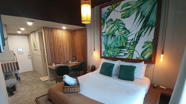 Une chambre d’hôtel avec un lit, un bureau et une chaise. Il y a une grande fenêtre avec des rideaux, un tableau représentant des feuilles accroché au mur et un luminaire suspendu.