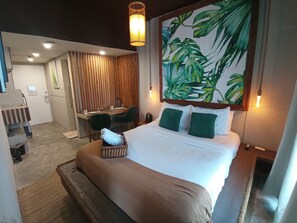 A hotel room with a bed, a desk, and a chair. There is a large window with curtains, a painting of leaves on the wall, and a hanging light fixture. - The Staycation - Boutique rooms (Makati)