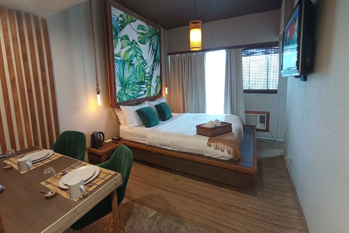 A modern hotel room with a large bed, a dining table set for two, and a large window with curtains.