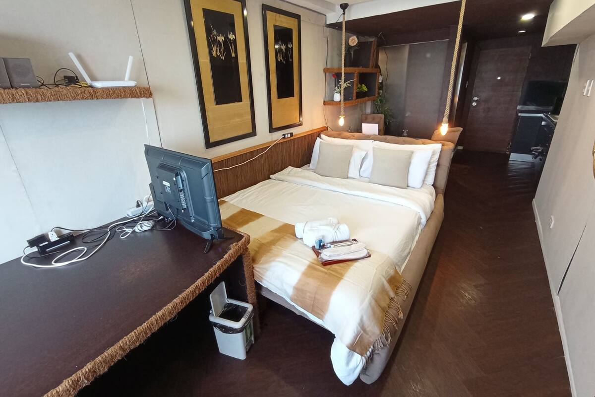 A hotel room with a bed, a desk with a computer, a TV, and a small trash bin.