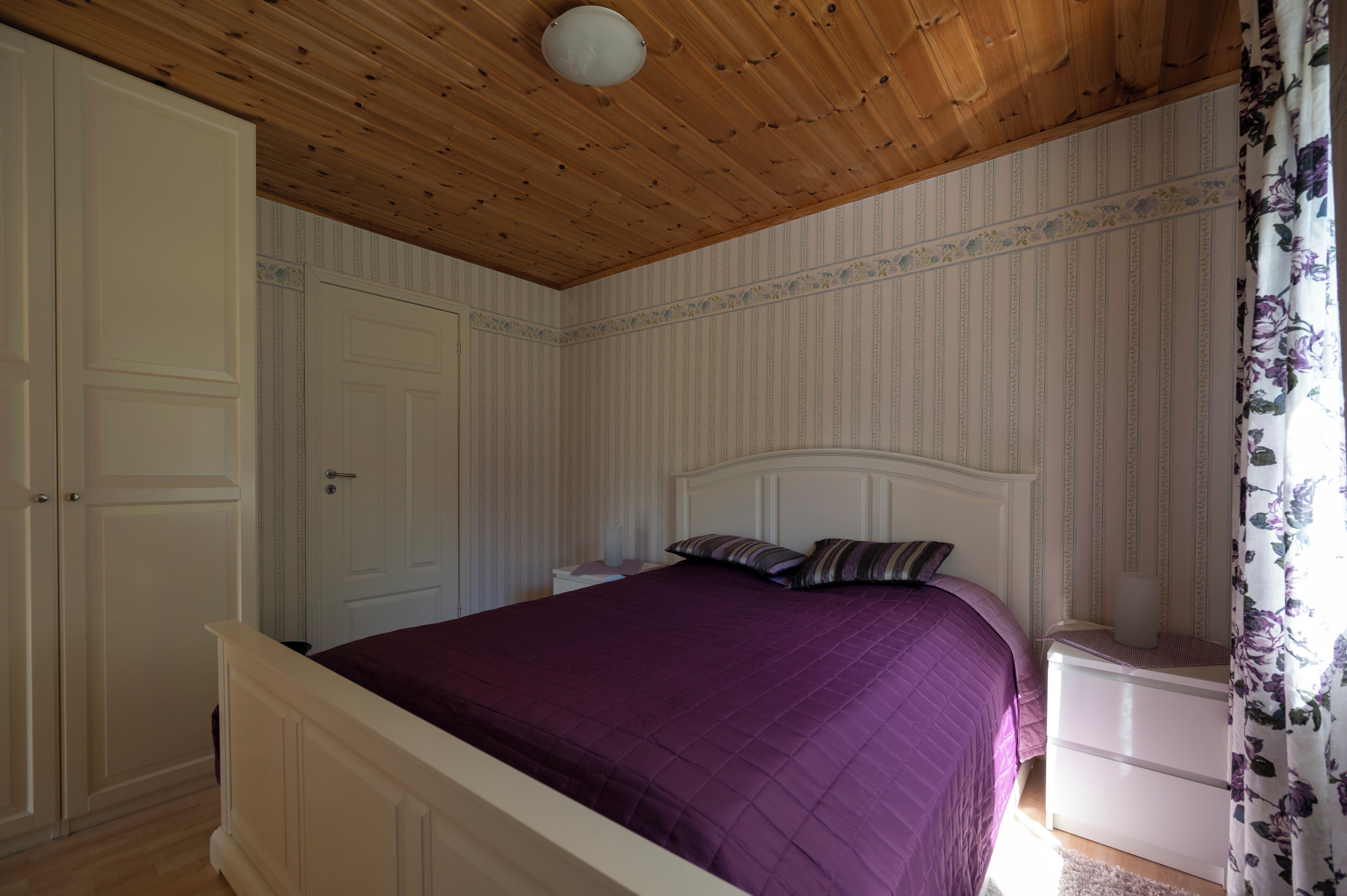 A bedroom with a wooden ceiling, a bed with a purple bedspread, a nightstand, and a white wardrobe.