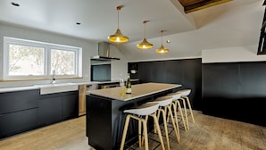 A modern kitchen with a large island, bar stools, pendant lighting, and a large window. - Le Chalets (Falls Creek)