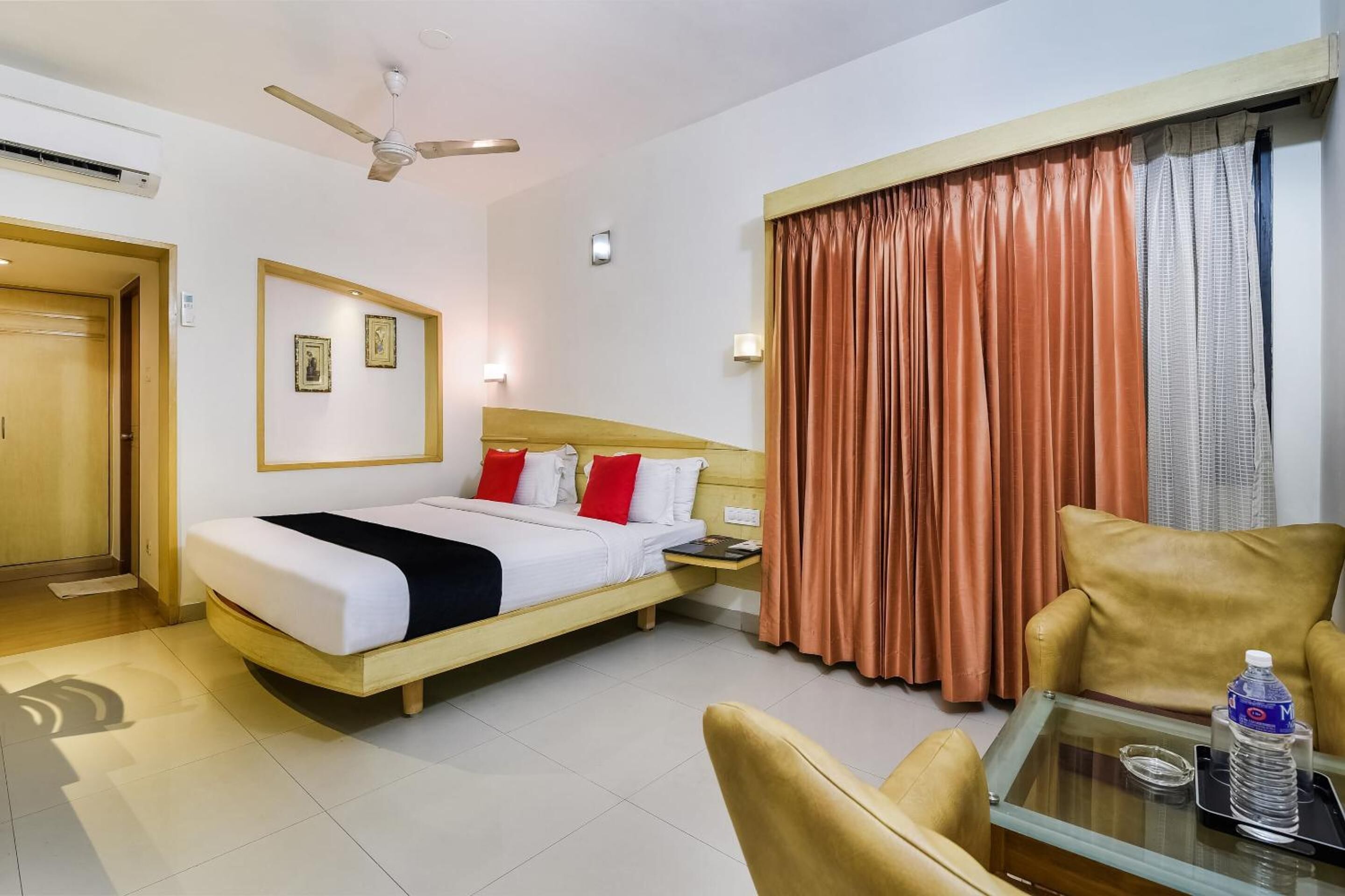 A hotel room with a bed, a chair, a table with a bottle, and a window with curtains.