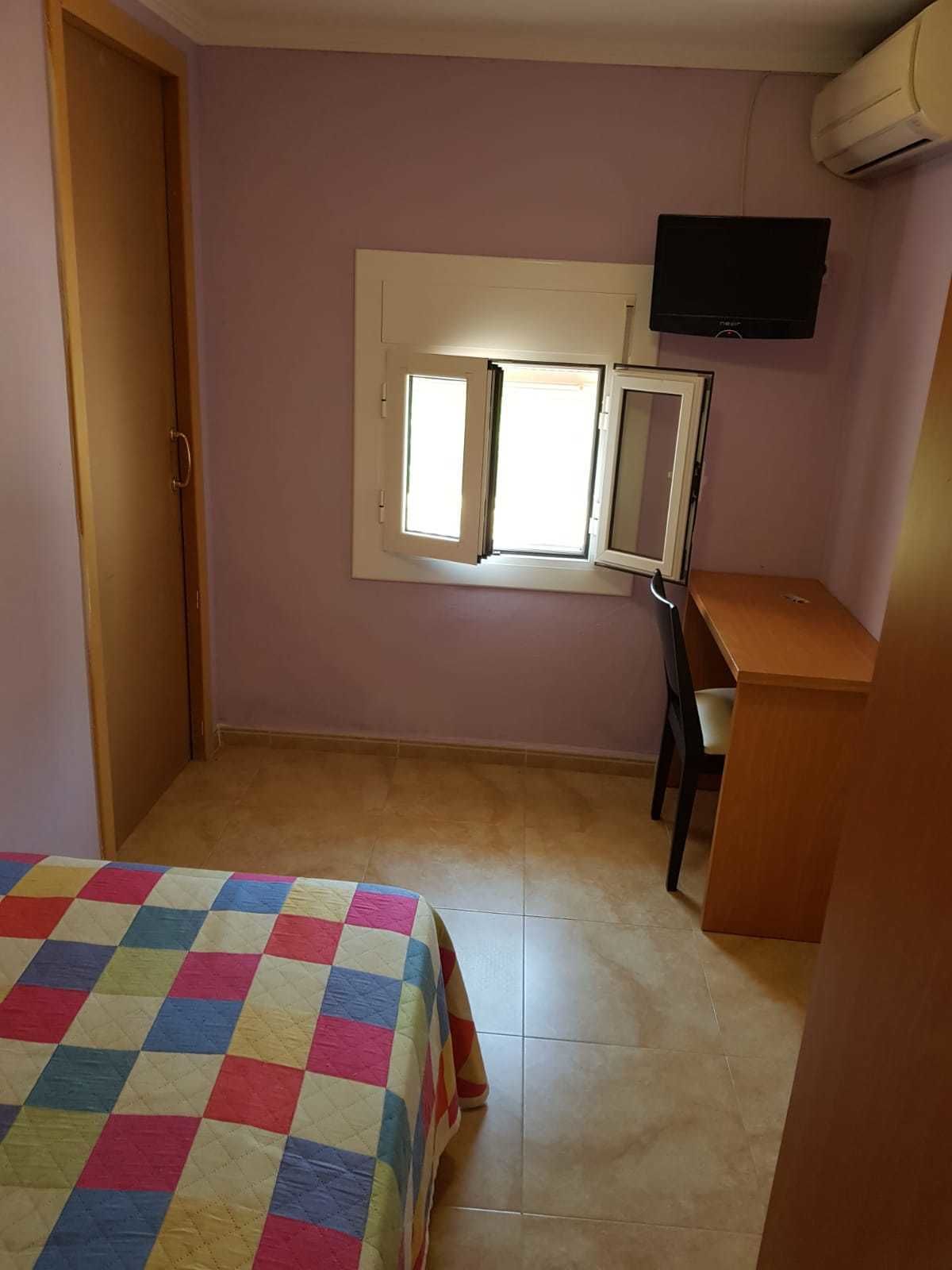 A bedroom with a bed, a desk, a chair, a television, and a window with blinds.