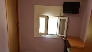A bedroom with a bed, a desk, a chair, a television, and a window with blinds.
