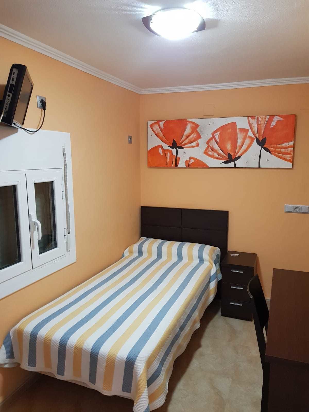 Een slaapkamer met een bed, een bureau, een stoel, een raam met jaloezieën en een schilderij aan de muur.