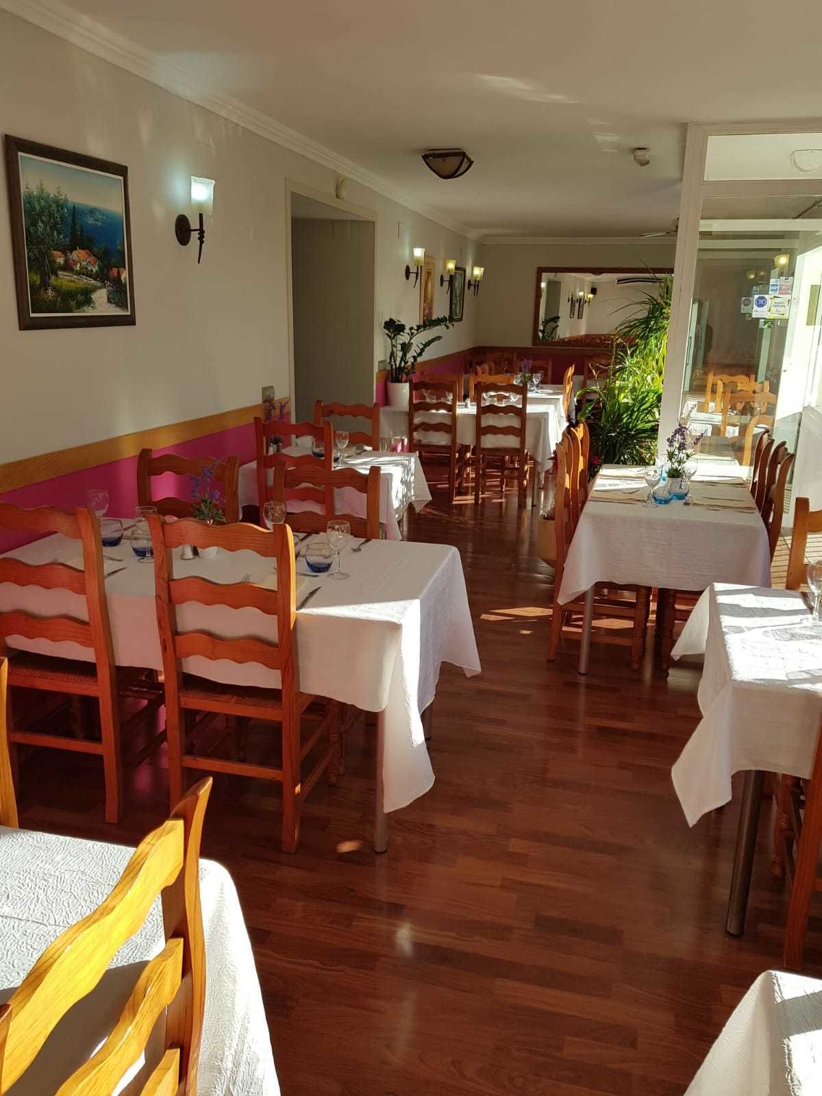 A dining area with wooden chairs and tables, a painting on the wall, and a potted plant.