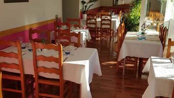 A dining area with wooden chairs and tables, a painting on the wall, and a potted plant.