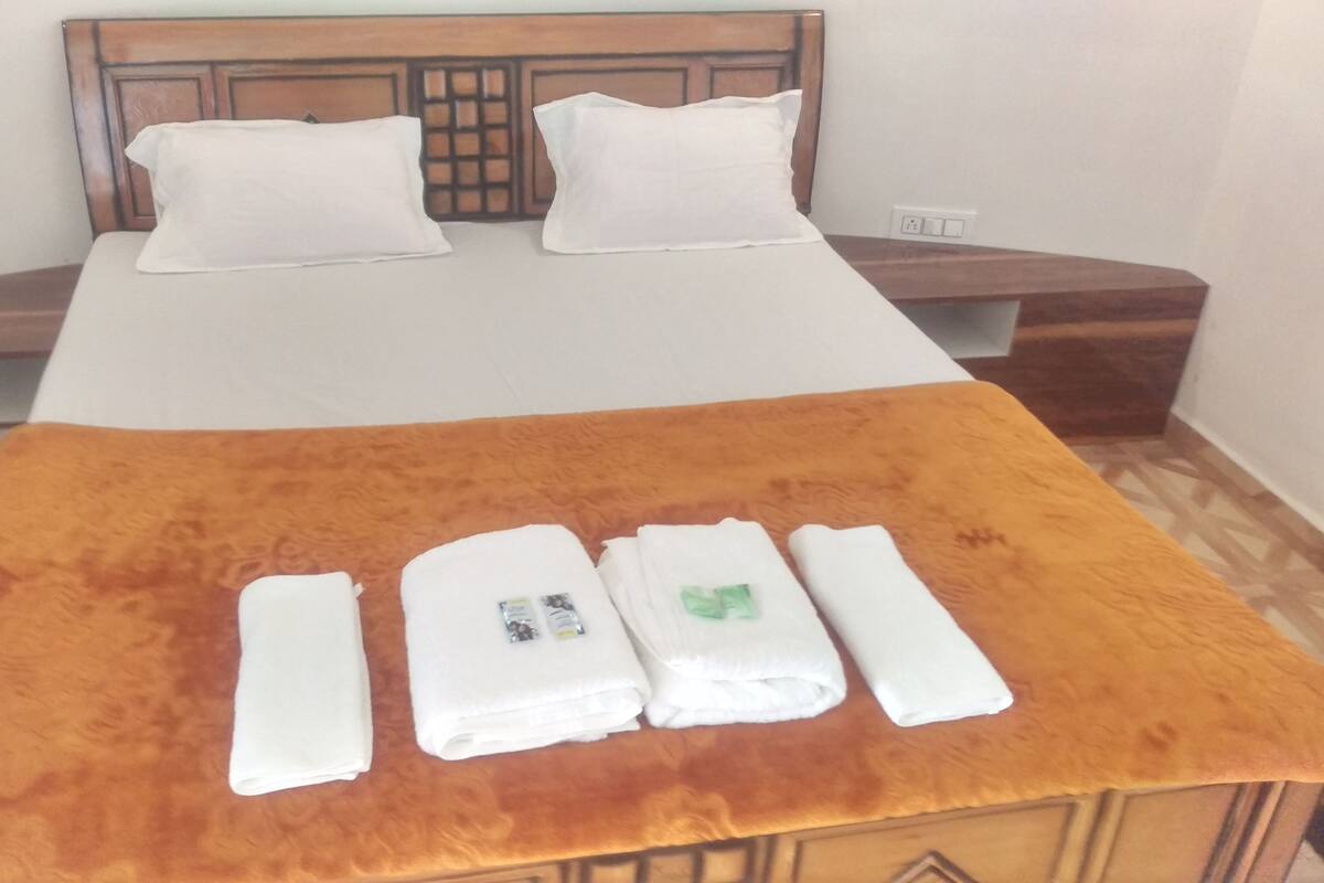 A hotel room with a bed, two pillows, a wooden headboard, a nightstand, and a table with towels and toiletries.