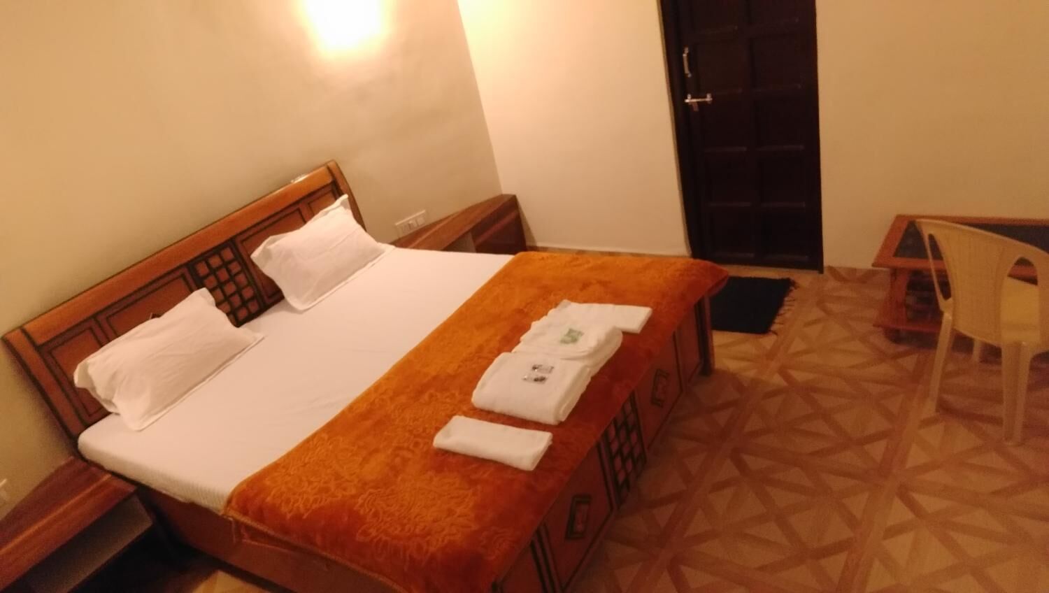 A neatly made bed with white linens and an orange blanket, a wooden headboard, a small bedside table, and a chair in a room with patterned flooring and a dark door.