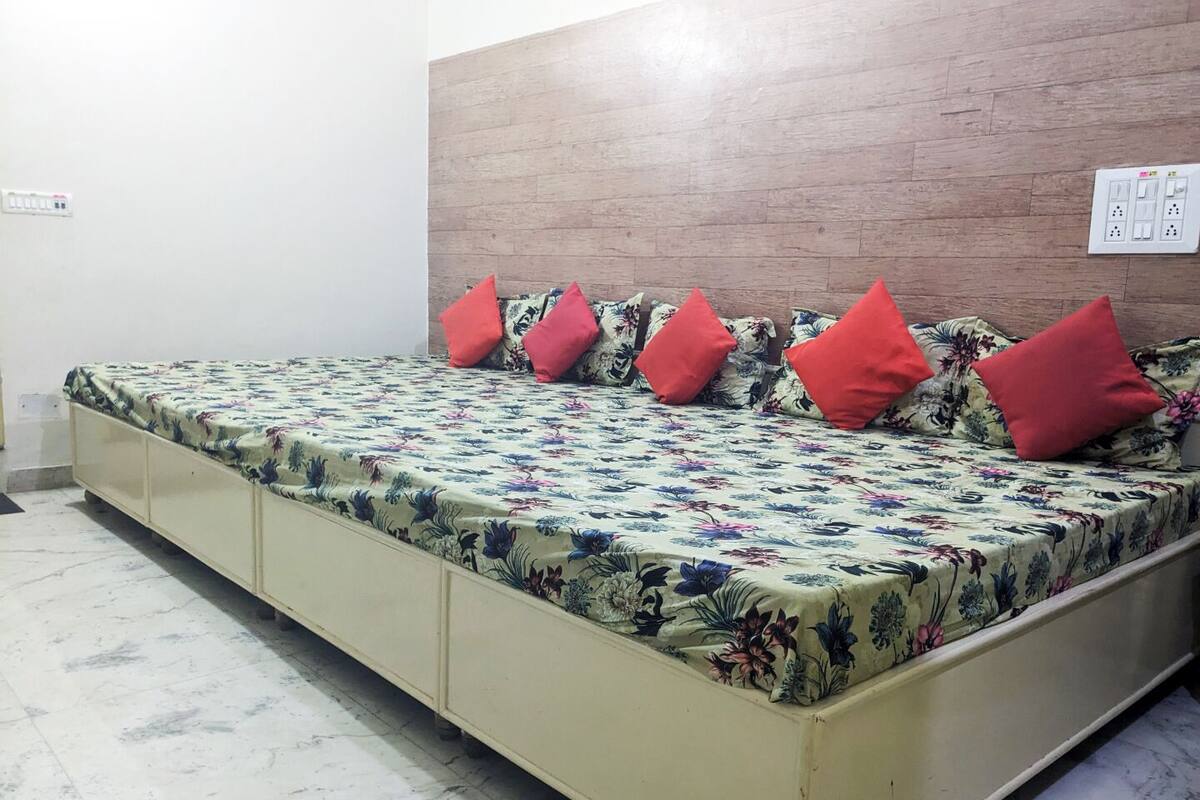 A single bed with a floral bedspread and red pillows, a wooden headboard, and a white electrical outlet on the wall.