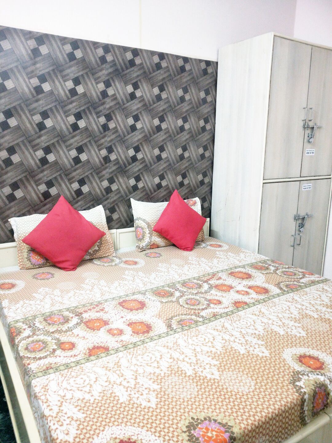 Una cama con colcha estampada y dos almohadas rojas, una pared a cuadros y un armario blanco de dos puertas.