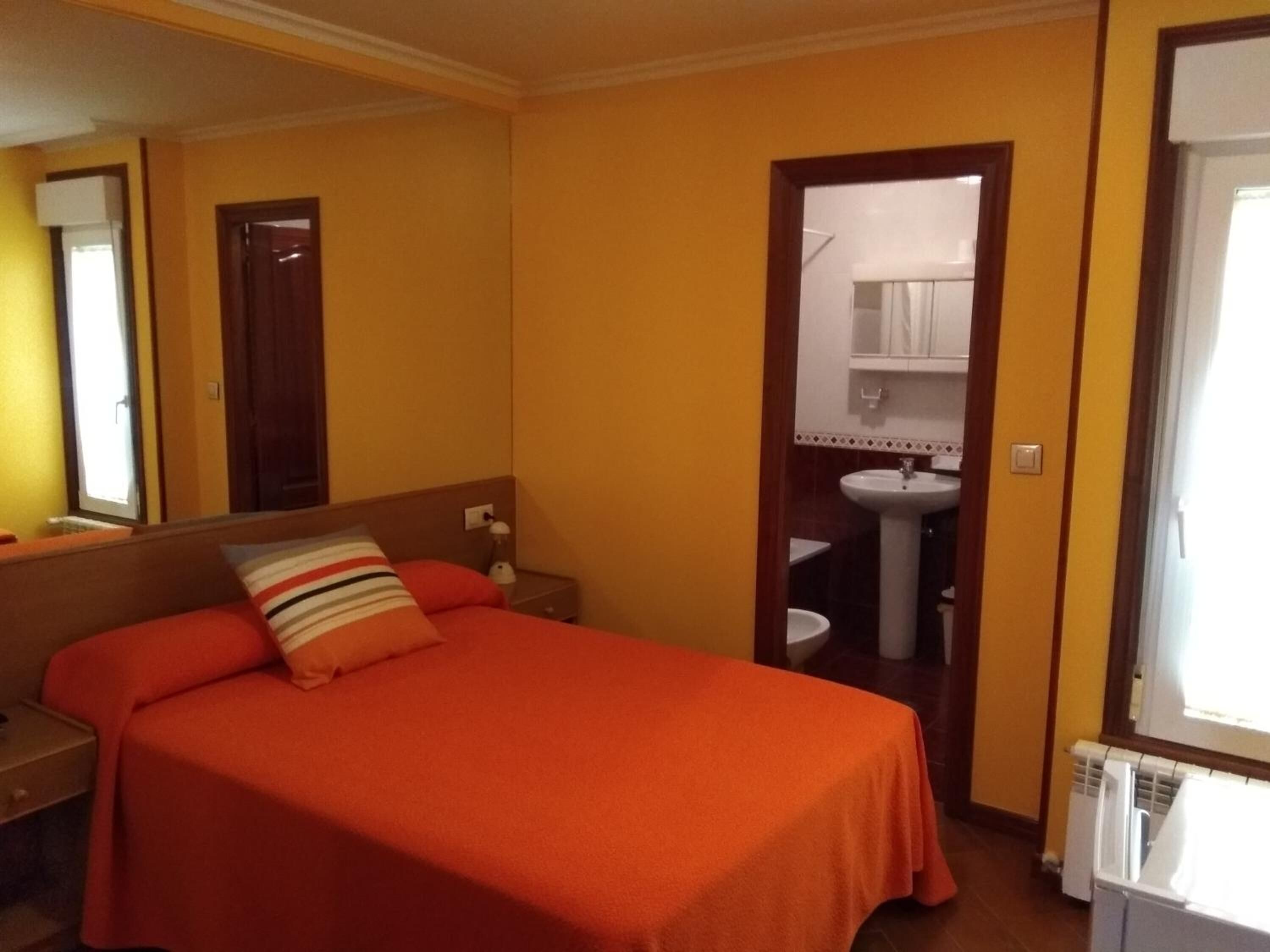 Habitación de hotel con una cama individual, una mesita de noche, un espejo y un baño con lavamanos e inodoro visibles a través de una puerta abierta.