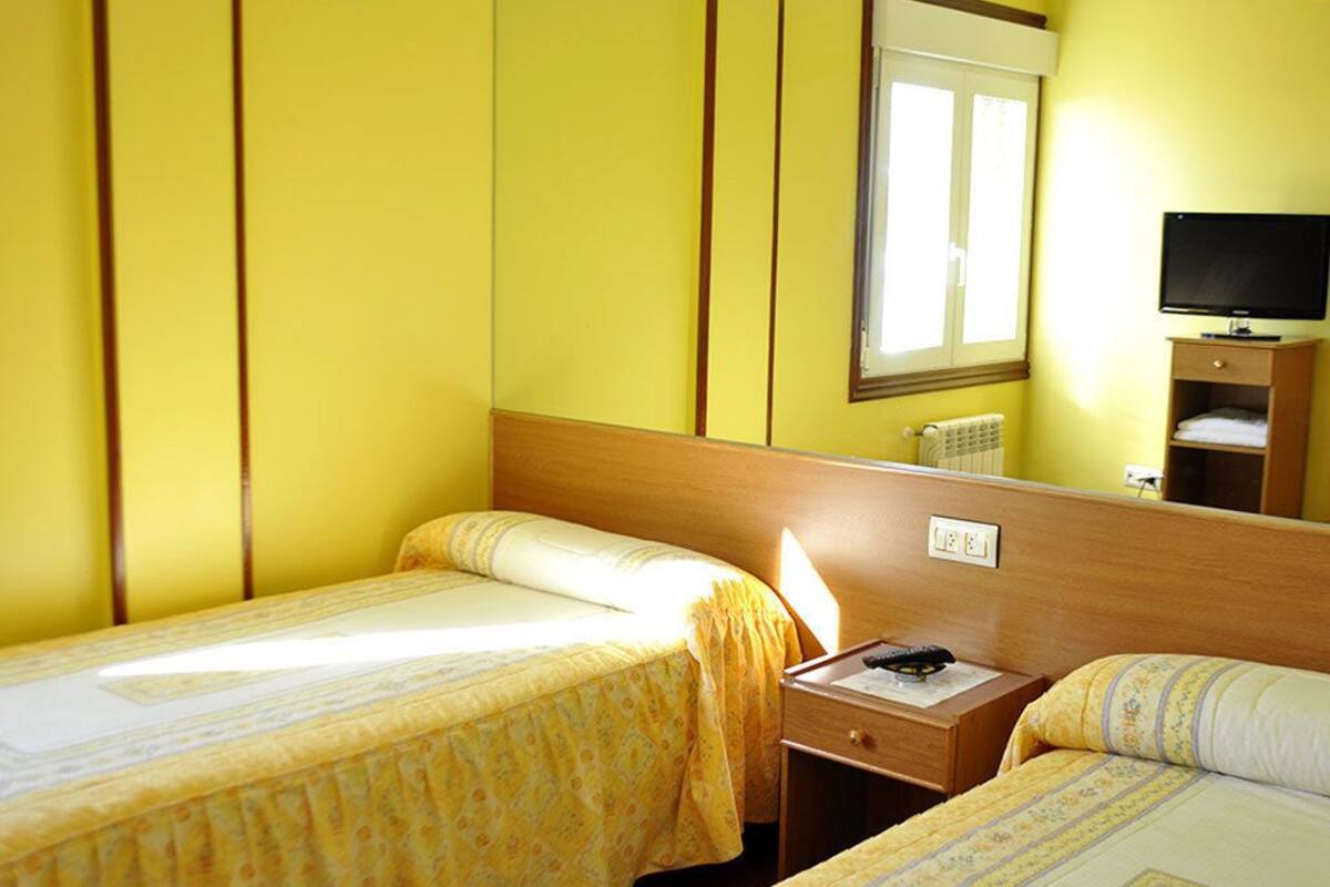 Habitación de hotel con dos camas individuales, cabecera de madera, mesita de noche, televisor y espejo.