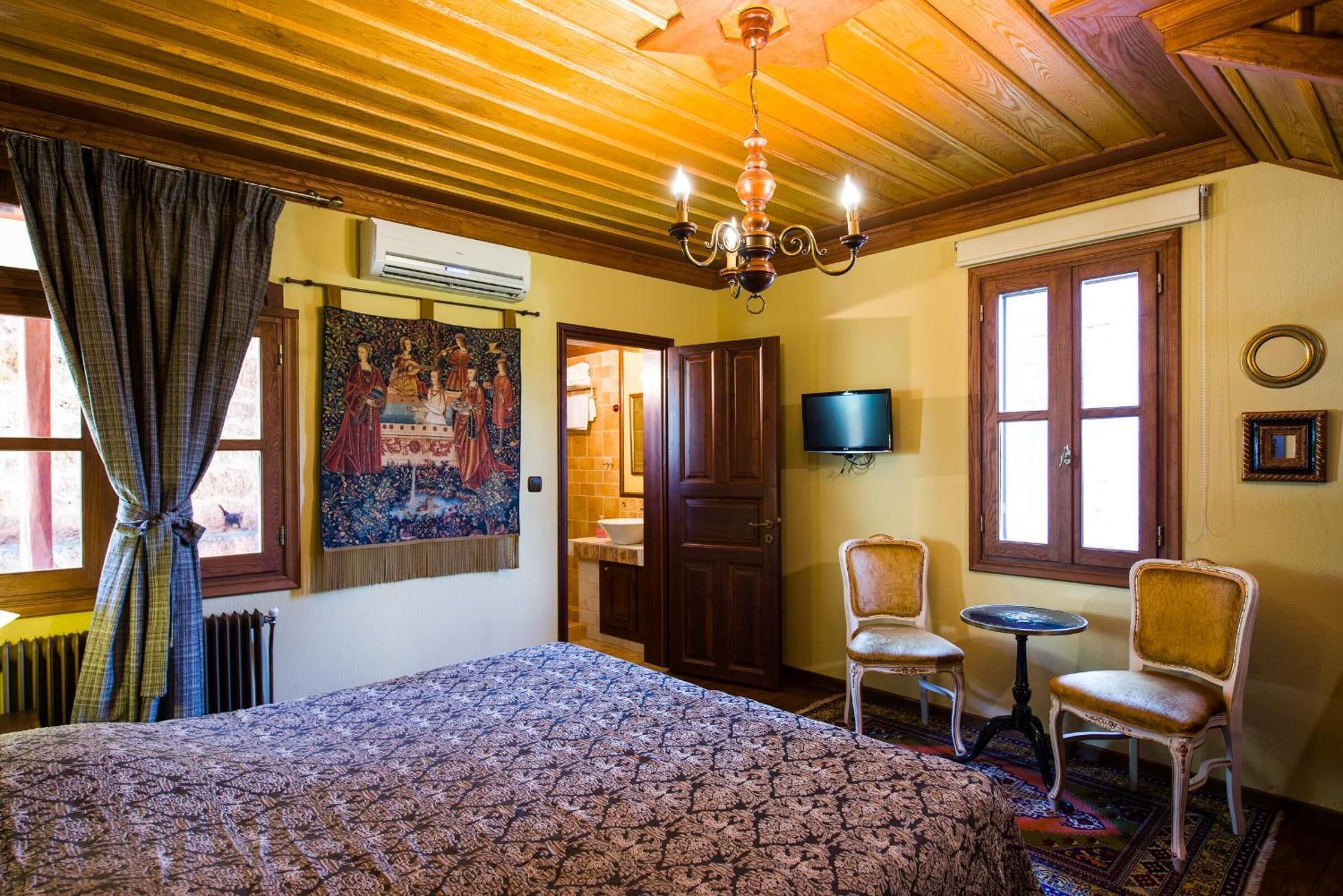 A room with a large bed, two chairs, a small table, a TV, and a wooden ceiling.
