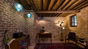 A rustic room with exposed wooden beams, a brick wall, a desk with a lamp, a chair, and a doll on a shelf.