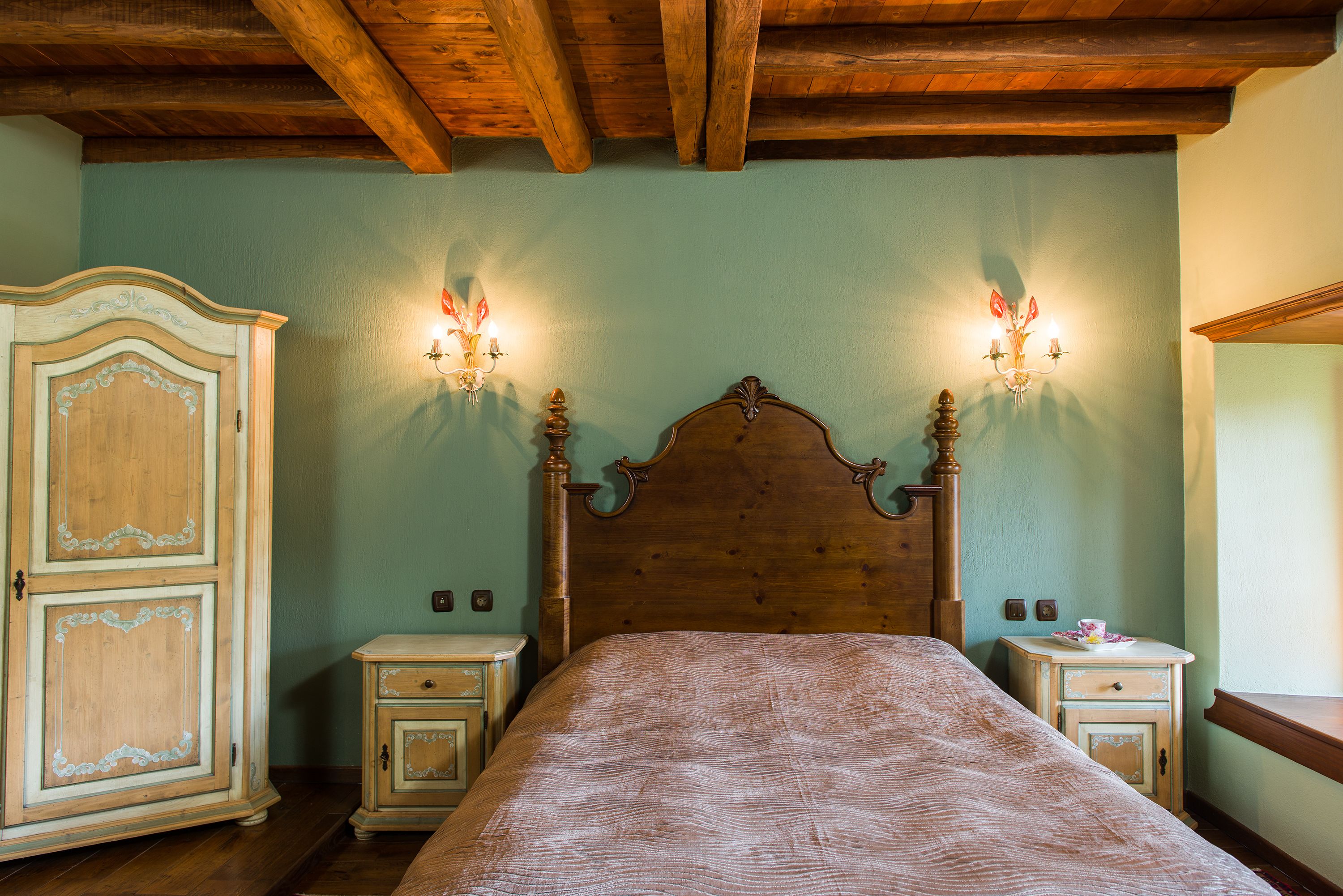 A bedroom with a wooden headboard, two bedside tables, a wardrobe, and wall sconces.