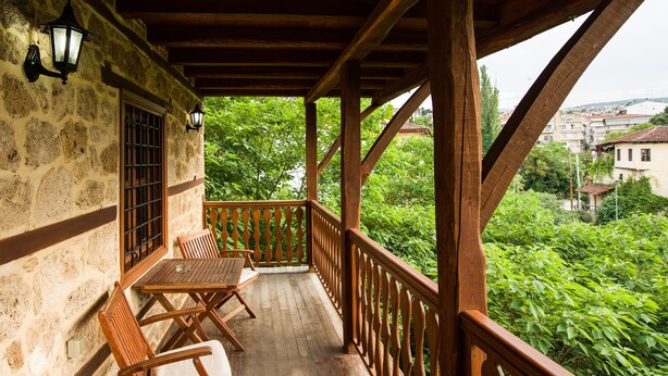 A wooden balcony with a table and chairs, stone walls, and a view of greenery and buildings in the background.