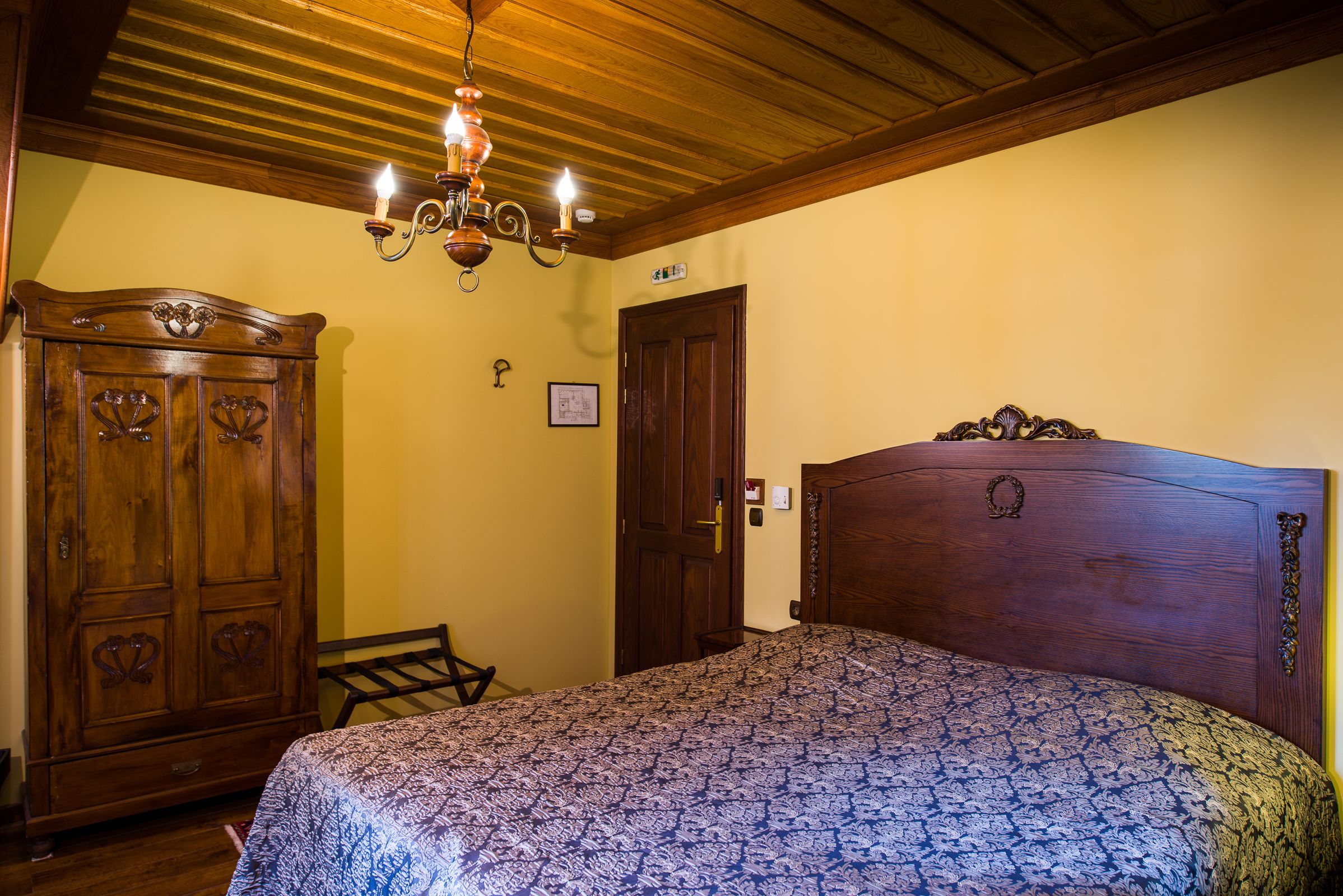 A bedroom with a wooden headboard, a patterned bedspread, a wooden wardrobe, and a chandelier.