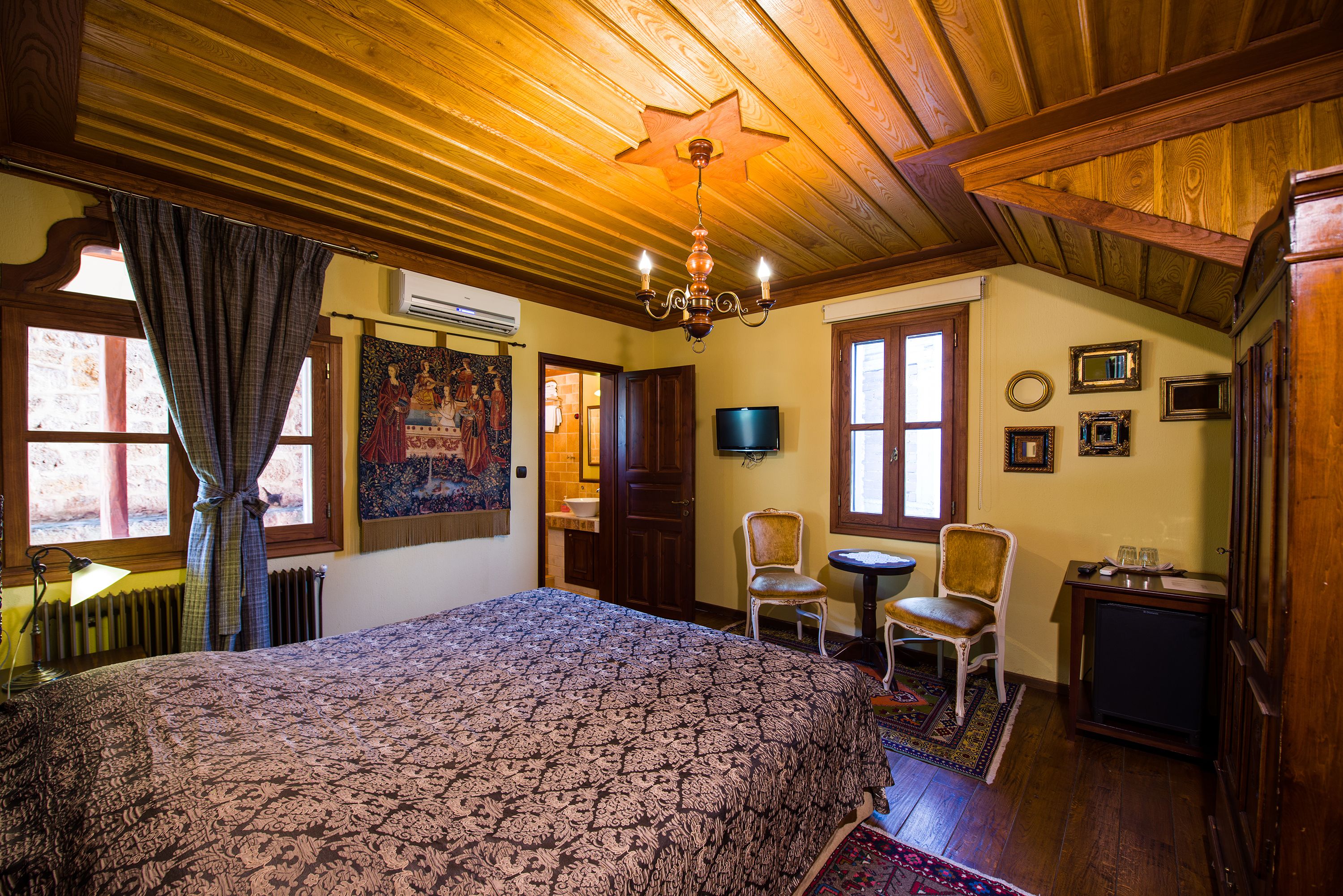 A room with a large bed, wooden ceiling, a chandelier, a television, and a small table with chairs.