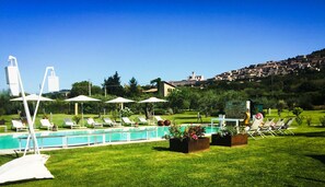 A pool area with lounge chairs and umbrellas, surrounded by a well-maintained lawn and a view of a hillside town.