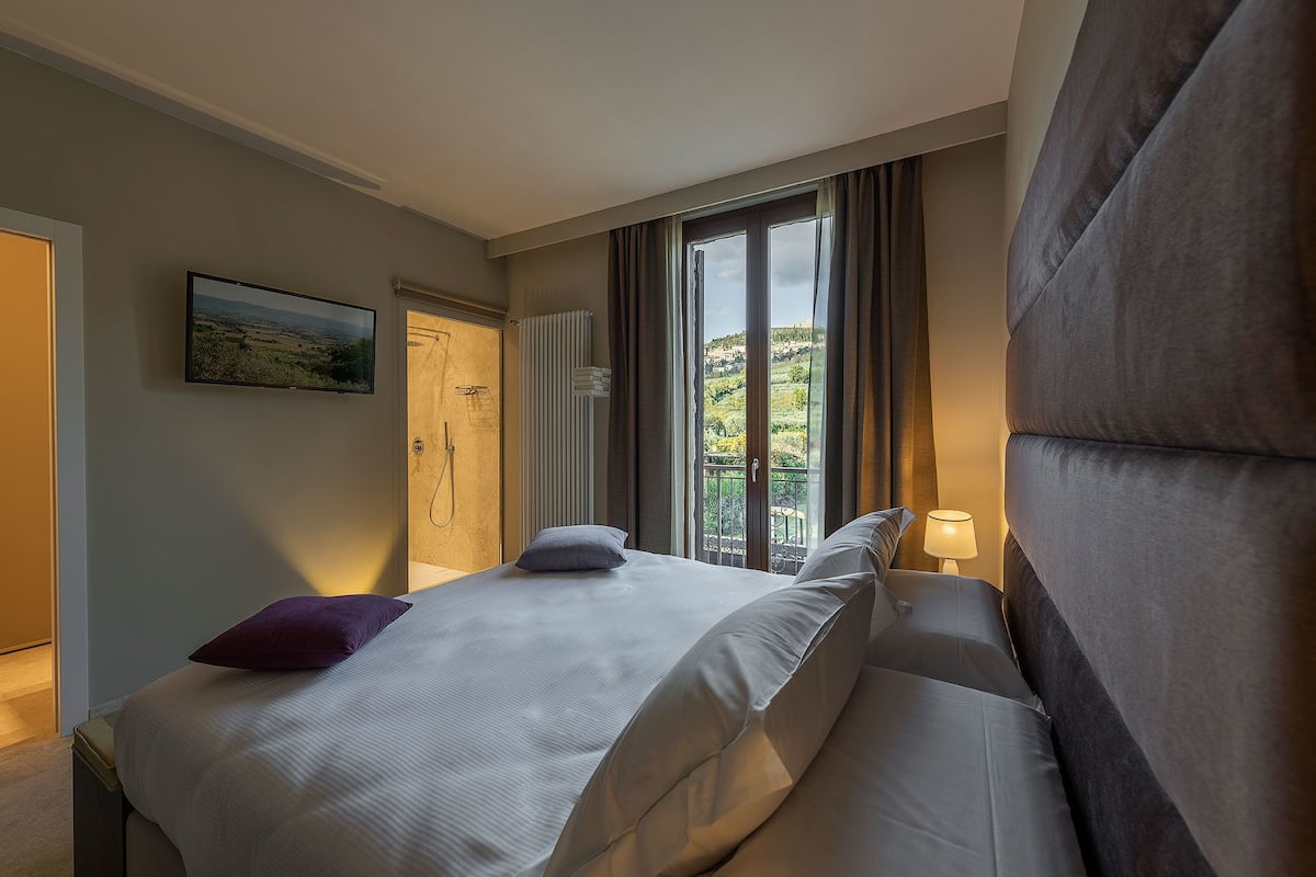 Hotellihuone, jossa on sänky, ikkuna, josta on näkymä vehreään maisemaan, ovi ja seinälle asennettu televisio.