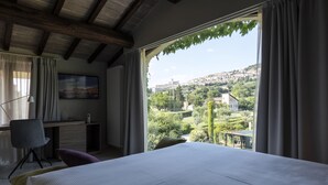 A bedroom with a large bed, a desk, and a chair. It has a view of a cityscape through a window. - Tenuta San Masseo (Assisi)