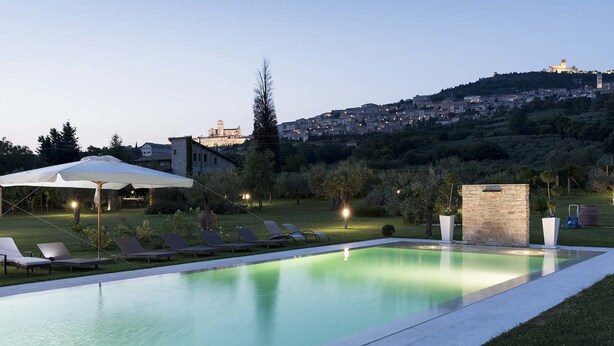 A swimming pool with lounge chairs and a stone structure, set against a backdrop of a hill with buildings. - Tenuta San Masseo (Assisi)