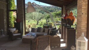A cozy outdoor seating area with wicker furniture, a large table, and a view of a lush garden and distant hills. - Tenuta San Masseo (Assisi)