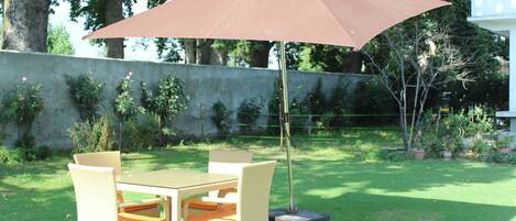 Um pátio externo com mesa e cadeiras, um guarda-sol castanho e uma parede de concreto com vegetação.
