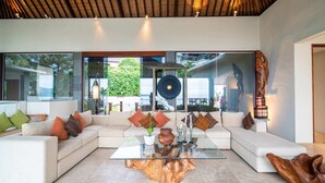A modern living room with a glass coffee table, white sofas, and a large window offering a view of greenery and a building. - Villa Gumamela Bali (Manggis)