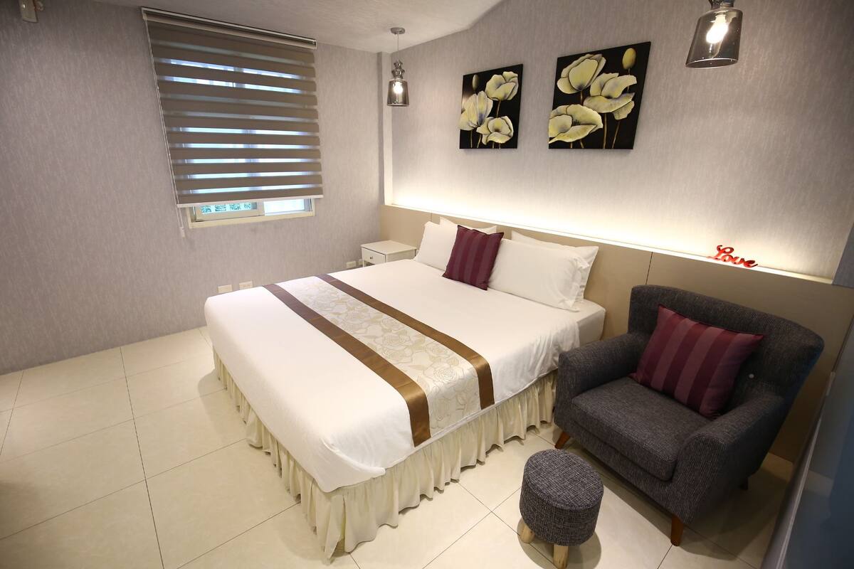Quarto com cama, cadeira, banquinho pequeno, mesa de cabeceira, luminária na parede e janela com persianas.