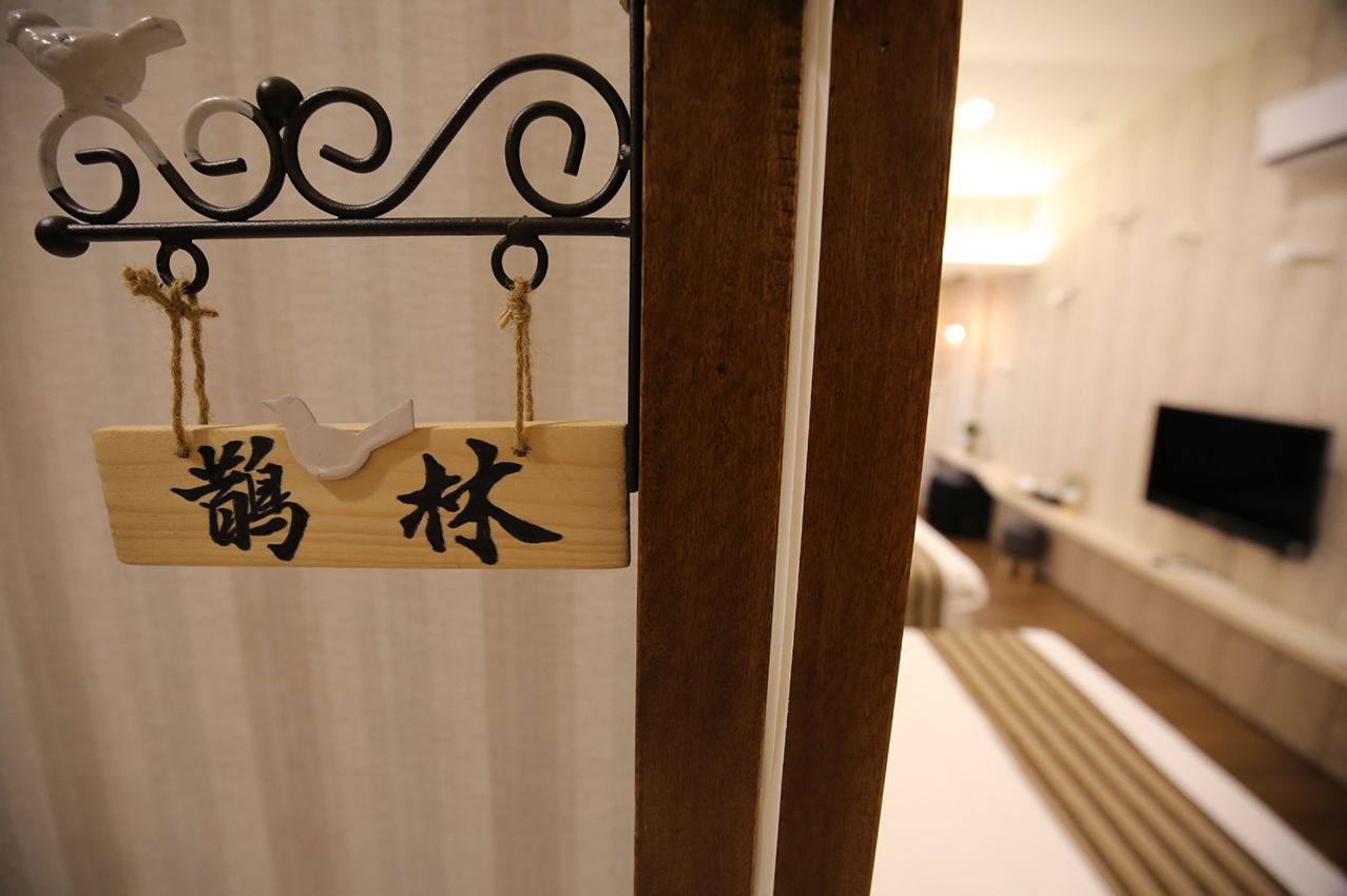 A wooden sign with Japanese characters, a black metal decorative element above it, and a blurred background of a room with a bed and a television.