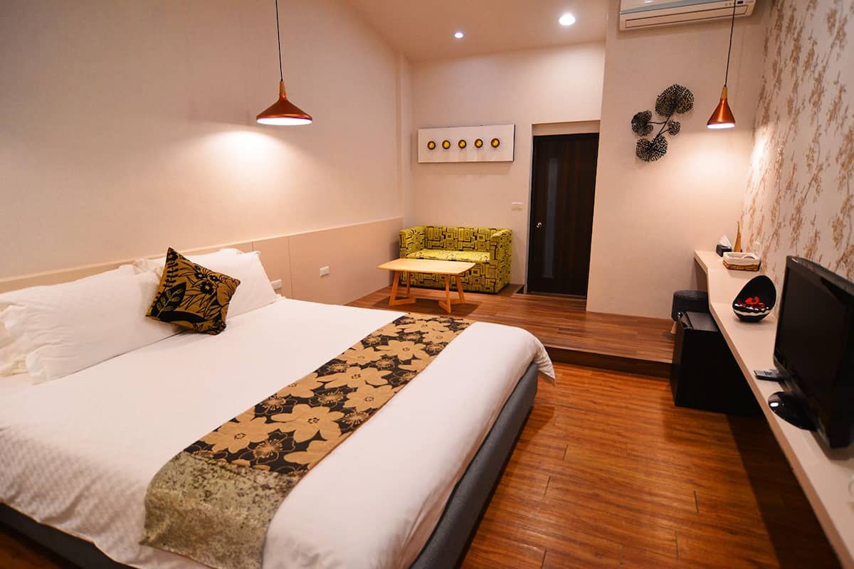 Quarto de hotel com cama, uma mesinha, televisão e um espaço de estar com uma poltrona estampada.